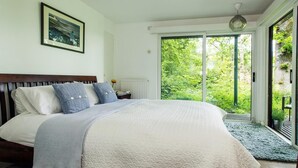 A bedroom with a large bed, a wooden headboard, a framed picture on the wall, and a view of greenery through a sliding glass door. - 2 Bed in Beaulieu (oc-nfl71) (Beaulieu)