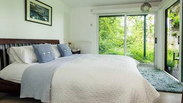 A bedroom with a large bed, a wooden headboard, a framed picture on the wall, and a view of greenery through a sliding glass door.