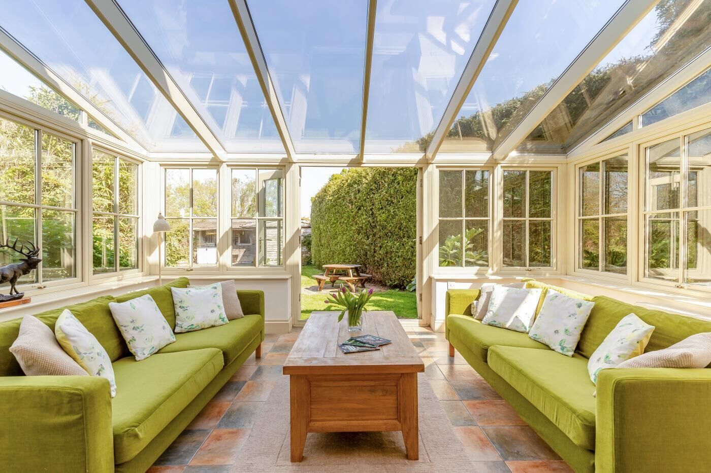 A conservatory with two green sofas, a wooden coffee table, and a view of a garden.