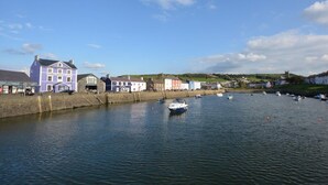 A waterfront hotel with a covered walkway, boats in the water, and a town in the background. - 2 Bed in Aberaeron (oc-45333) (Aberaeron)
