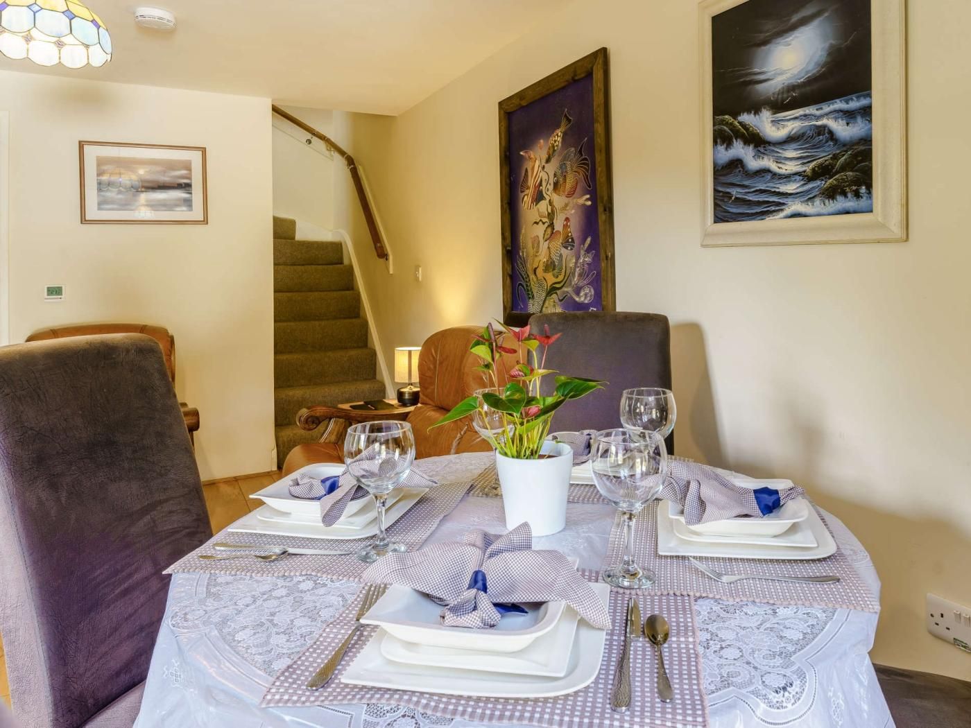 A dining area with a table set for four, a staircase in the background, and a painting on the wall.