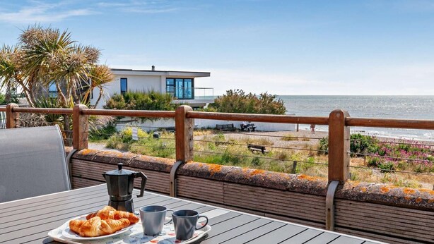 A terrace with a table set for breakfast, overlooking a beach and a modern building. - 4 Bed in Pevensey Bay (oc-bt069) (Pevensey Bay)