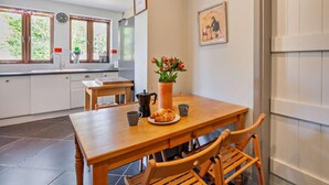A modern kitchen with a dining area, featuring a wooden table, chairs, a vase with flowers, and a clock on the wall. - 4 Bed in Pevensey Bay (oc-bt069) (Pevensey Bay)