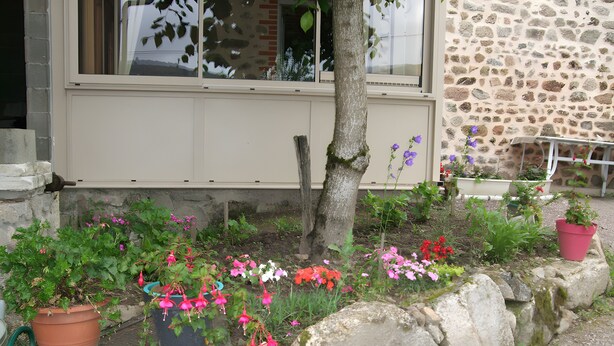 A garden with potted plants and flowers, a stone wall, and a building with windows. - The "Grand Reu" Cottage - Gite Camille (Corancy)