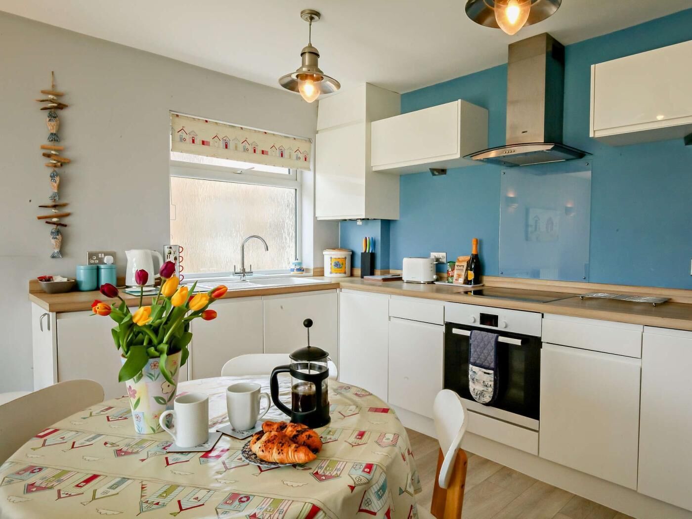 A kitchen with white cabinets, a blue wall, a window, and a table with a tablecloth, coffee pot, and croissants.