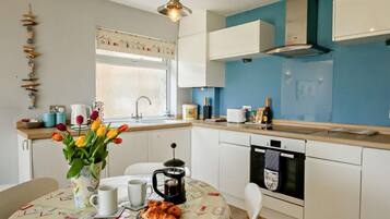 A kitchen with white cabinets, a blue wall, a window, and a table with a tablecloth, coffee pot, and croissants.