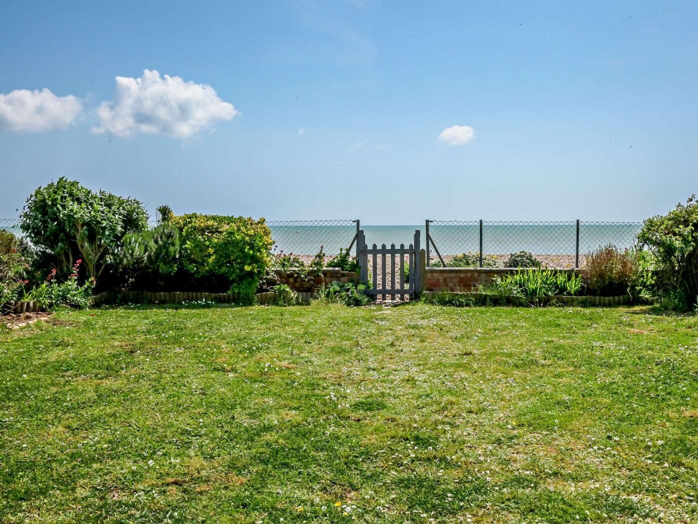 A grassy area with a gate leading to a beach, surrounded by greenery and a fence.