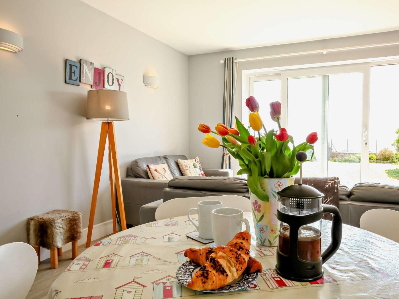 Un accogliente soggiorno con un divano, un tavolino con croissant e una caffettiera, un vaso con tulipani e una lampada.