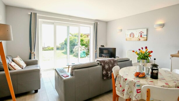 Ein Wohnzimmer mit grauem Sofa, einem Fernseher, einem Esstisch mit Stühlen und einem großen Fenster mit Vorhängen.