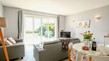 A living room with a grey sofa, a TV, a dining table with chairs, and a large window with curtains.