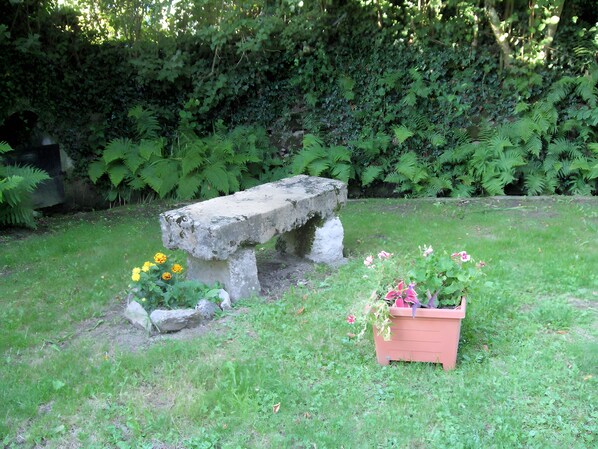 A stone bench in a garden with grass and ferns, a planter with flowers, and a dense green hedge in the background.
