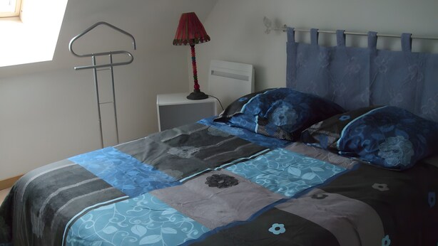 A bed with a blue and gray quilt, a red lamp, and a window with a curtain. - Le gîte du "Grand Reu" - Gîte Anna (Corancy)