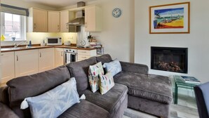 A modern kitchen with beige cabinets, a microwave, and a stove. There is a gray sofa with colorful pillows and a fireplace with a framed painting above it. - 2 Bed in Camber (oc-bt084) (Camber)