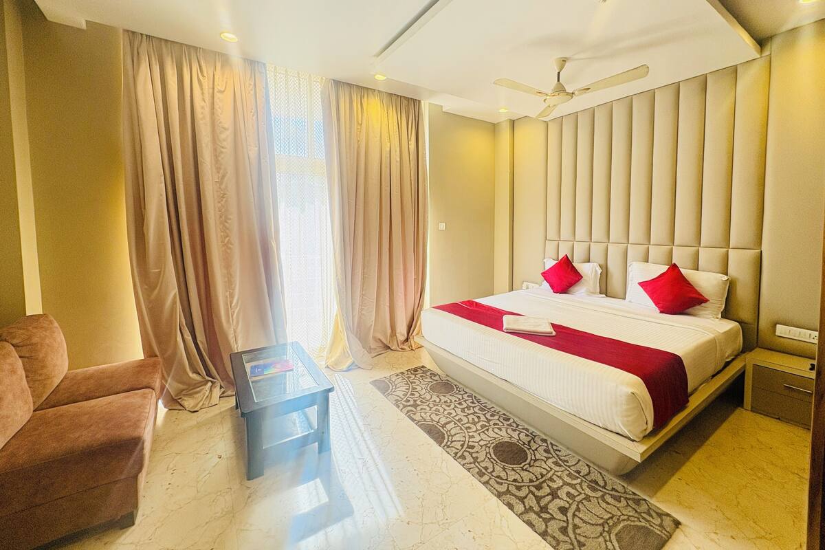 Una habitación de hotel con cama, una silla, una mesita, ventilador de techo y ventana con cortinas.