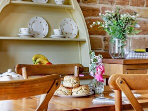 A wooden dining table with a plate of scones, a vase with flowers, and a brick wall in the background.