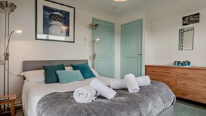 A neatly made bed with a grey blanket, teal pillows, and rolled white towels. There is a wooden dresser with a mirror, a framed picture on the wall, and a floor lamp. - 1 Bed in Penzance (oc-47390) (Pendeen)