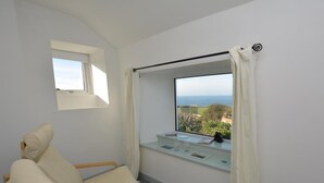 A room with a bed, a window with a view of the sea, and a small table with books and a cup. - 1 Bed in Penzance (oc-47390) (Pendeen)