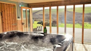 A hot tub with a bottle and glasses, a wooden door, and a view of a green landscape through large windows. - 4 Bed in Boscastle (oc-47035) (Forrabury and Minster)