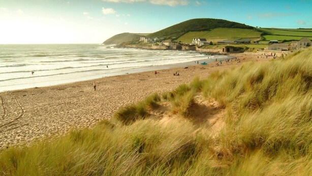 Une plage côtière bordée de dunes herbeuses, un ciel dégagé et, en arrière-plan, des collines parsemées de bâtiments.