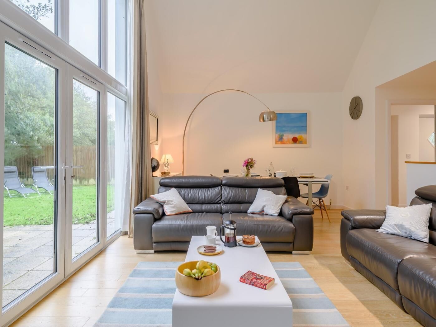 A modern living room with a sofa, a coffee table with fruits and books, a dining area with a table and chairs, and large windows.