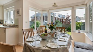 A dining area with a table set for four, a vase with flowers, and a view of houses through large windows. - 4 Bed in Rye- (oc-bt104) (Rye)