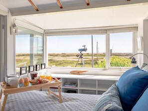 Ein Zimmer mit großem Fenster, einem Teleskop, einem Tablett mit Frühstücksgegenständen und einem Bett mit blauem Kopfkissen.