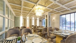 Un espace repas avec des tables dressées pour quatre personnes, des chaises, un lustre et de grandes fenêtres.