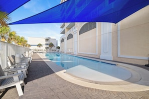 Une piscine dotée de chaises longues et d’un espace ombragé, entourée d’immeubles avec des balcons.