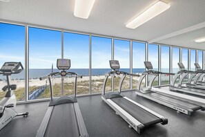 Une salle de sport équipée de plusieurs tapis de course et de vélos d’exercice, avec de grandes fenêtres donnant sur la plage.