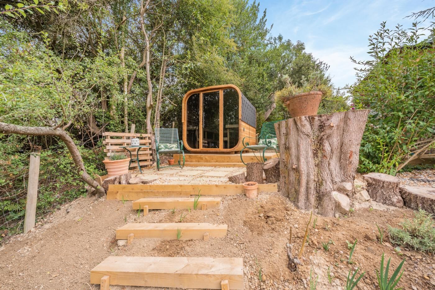 A wooden cabin with a large window, surrounded by a garden with steps leading to it.