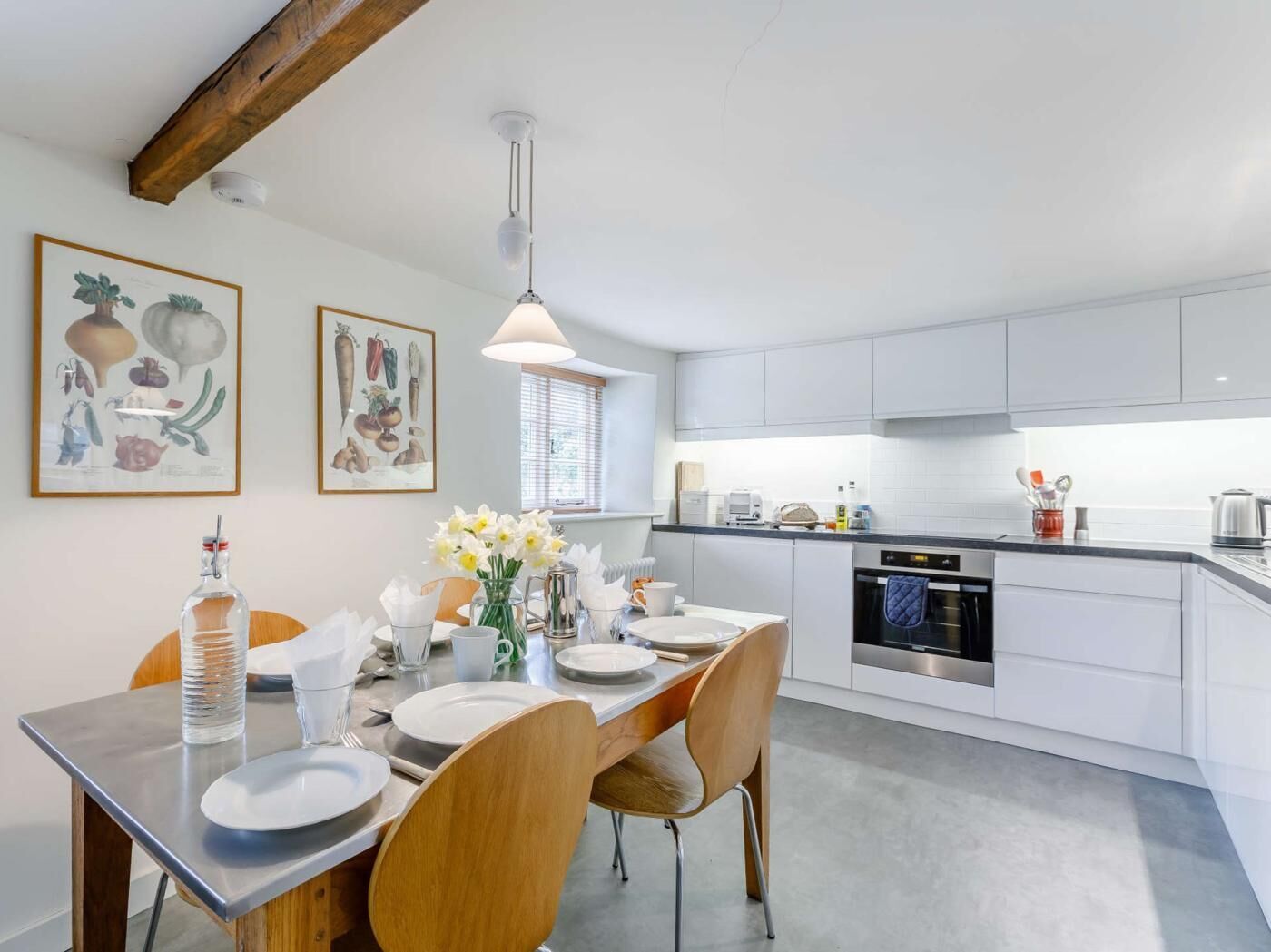 A modern kitchen with white cabinets, a dining table set with plates and a vase of flowers, and a hanging light fixture.