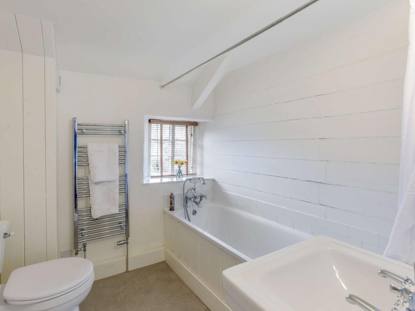 Une salle de bain avec une baignoire blanche, des toilettes, un porte-serviettes et une fenêtre avec des stores.