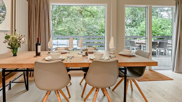 Ein Essbereich mit großem Tisch, gedeckt für vier Personen, Stühlen mit Holzbeinen, hängenden Pendelleuchten und Blick ins Grüne durch große Fenster.
