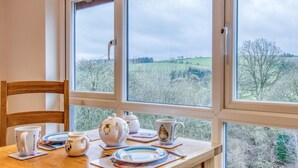 Ein Esstisch mit Tee-Service und Tellern, ein Holzstuhl und ein Fenster mit Blick auf eine grüne Landschaft.