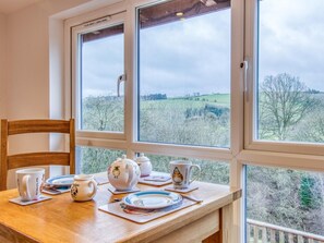 Ein Esstisch mit Tee-Service und Tellern, ein Holzstuhl und ein Fenster mit Blick auf eine grüne Landschaft.