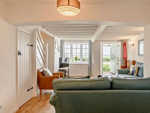 Ein geräumiges Wohnzimmer mit einem großen, grünen Sofa, einem Holztisch und einem Fernseher. Eine Tür führt auf einen Balkon mit Blick ins Grüne.