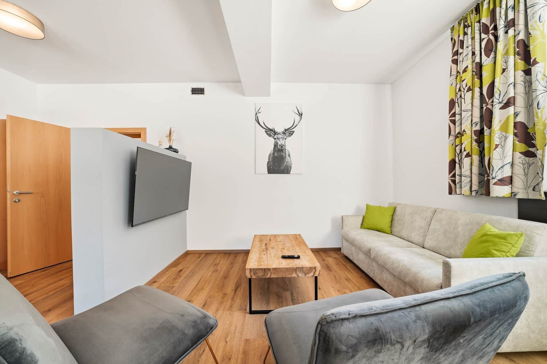 Ein modernes Wohnzimmer mit Fernseher, Couchtisch aus Holz, einer Couch mit grünen Kissen und einem Wandbild mit Hirschmotiven.