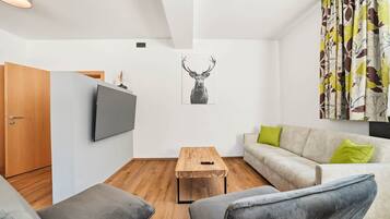 Ein modernes Wohnzimmer mit Fernseher, Couchtisch aus Holz, einer Couch mit grünen Kissen und einem Wandbild mit Hirschmotiven.