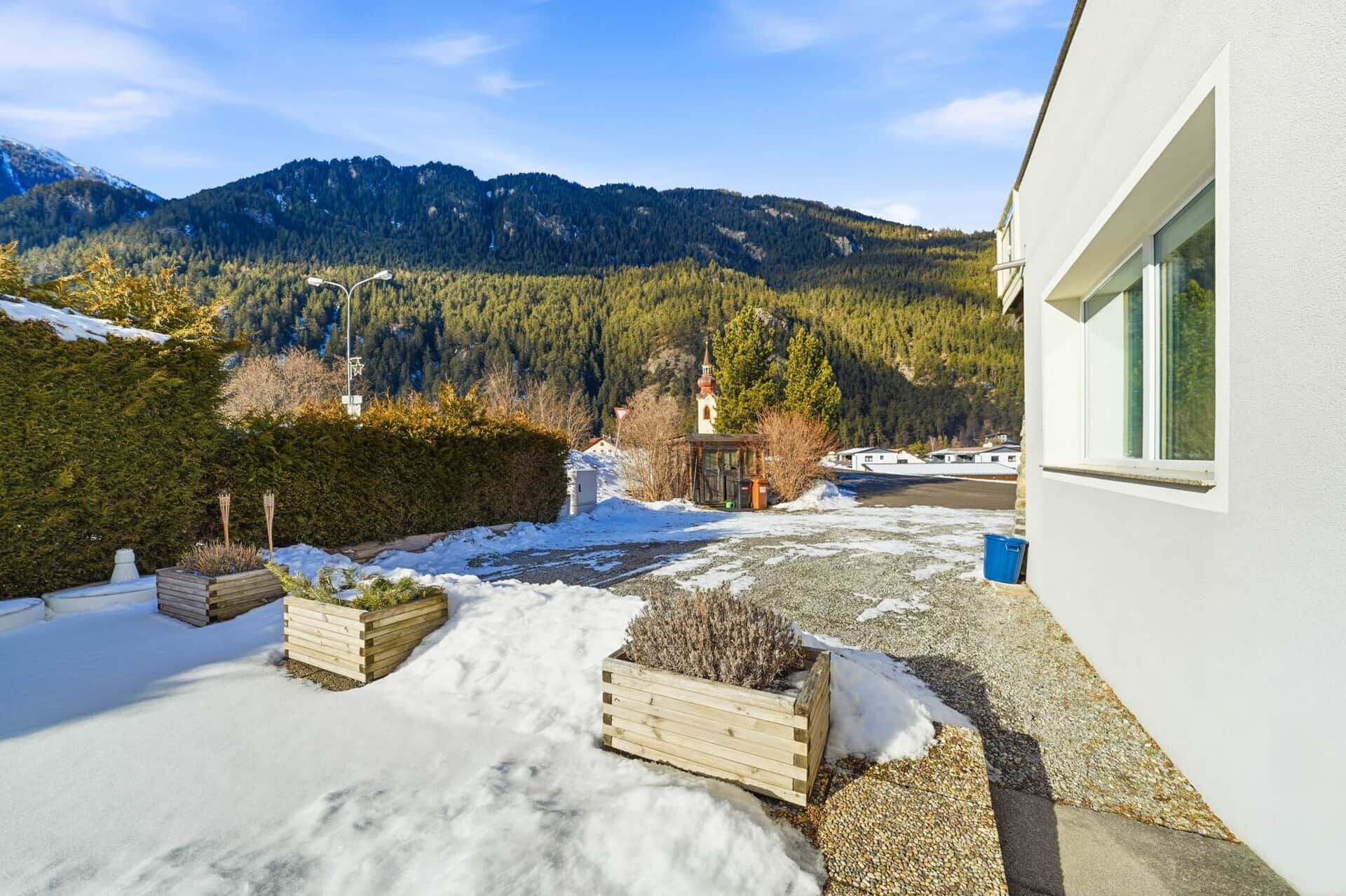 A snowy outdoor area with wooden planters, a small building, and a mountain backdrop.