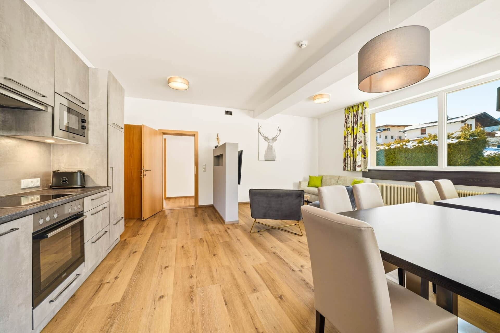 Eine moderne Küche mit integrierten Geräten, ein Essbereich mit großem Tisch und Stühlen sowie ein Blick ins Freie durch ein Fenster.