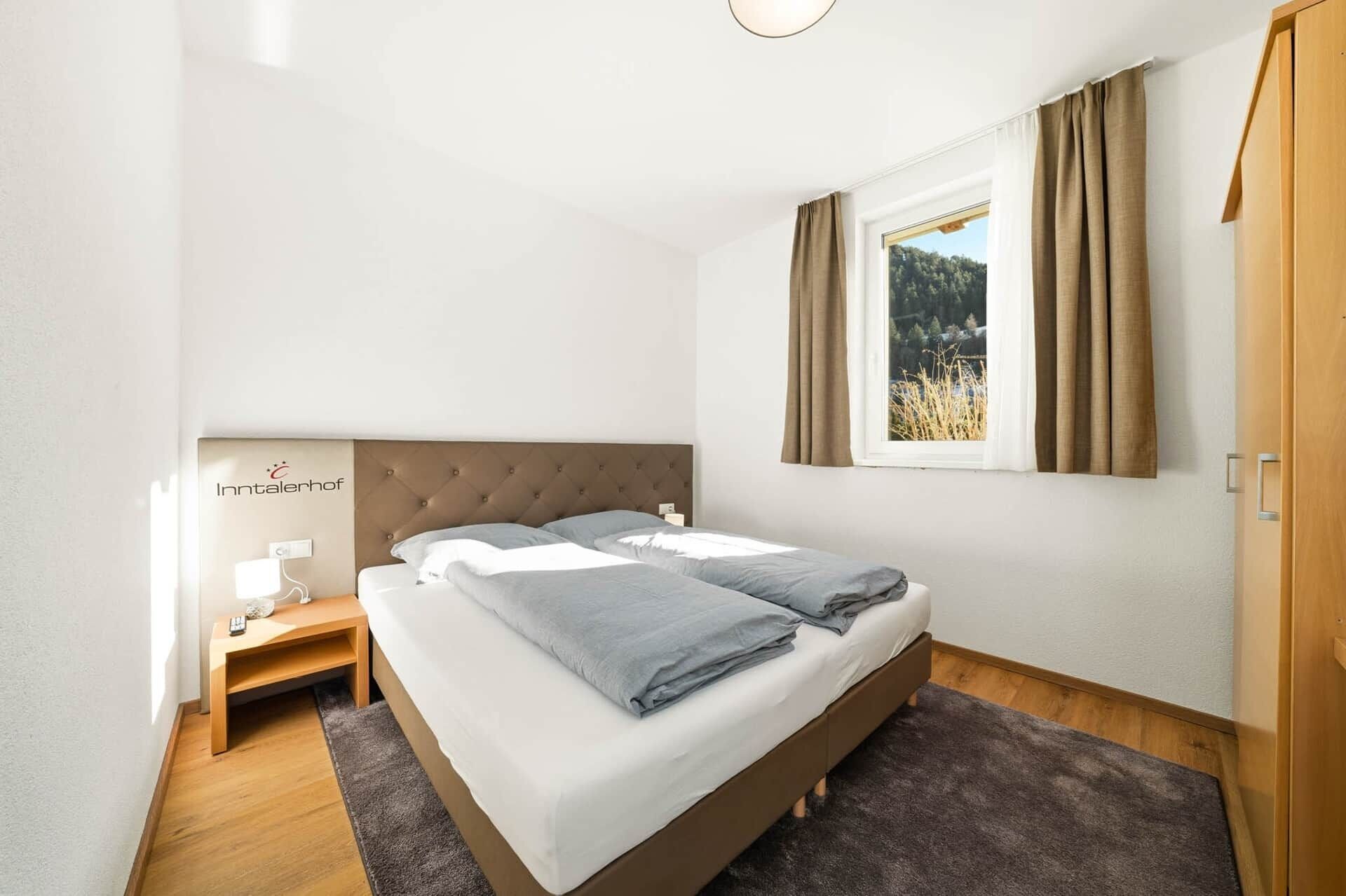 Ein Schlafzimmer mit Bett, Nachttisch, Lampe und Blick auf einen Wald durch ein Fenster.