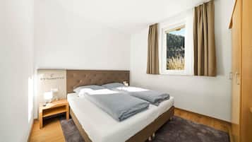 Ein Schlafzimmer mit Bett, Nachttisch, Lampe und Blick auf einen Wald durch ein Fenster.
