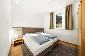 Un dormitorio con cama, mesita de noche, lámpara y vistas a un bosque por la ventana.