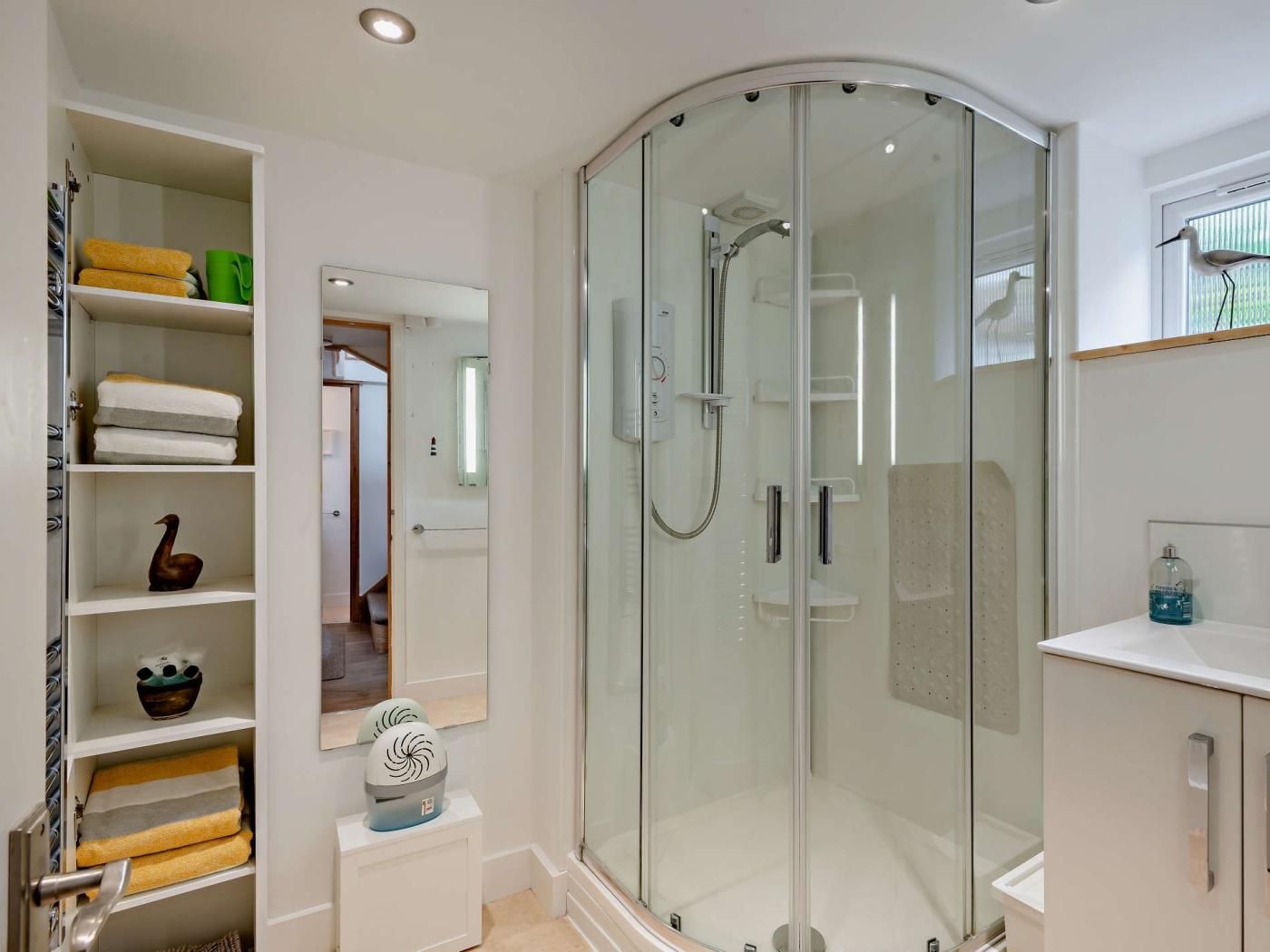 Ein modernes Badezimmer mit einer Glaskabine zur Dusche, einem Waschtisch mit Spiegel und einer integrierten Ablage mit Handtüchern und Dekoration.