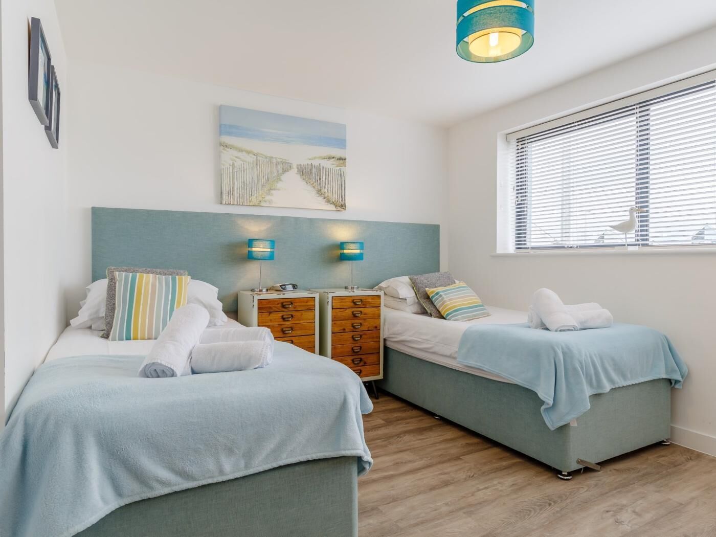A bedroom with two beds, a wooden dresser, bedside lamps, a painting, and a window with blinds.