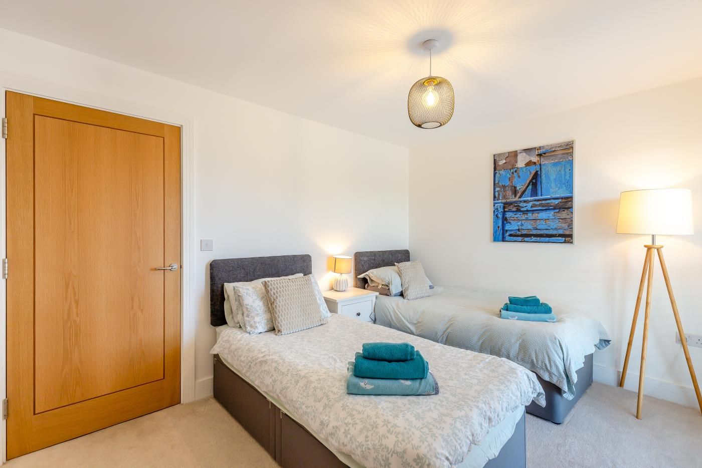 Un dormitorio con dos camas, una puerta de madera, un cuadro, una lámpara de pie y un aplique de techo.