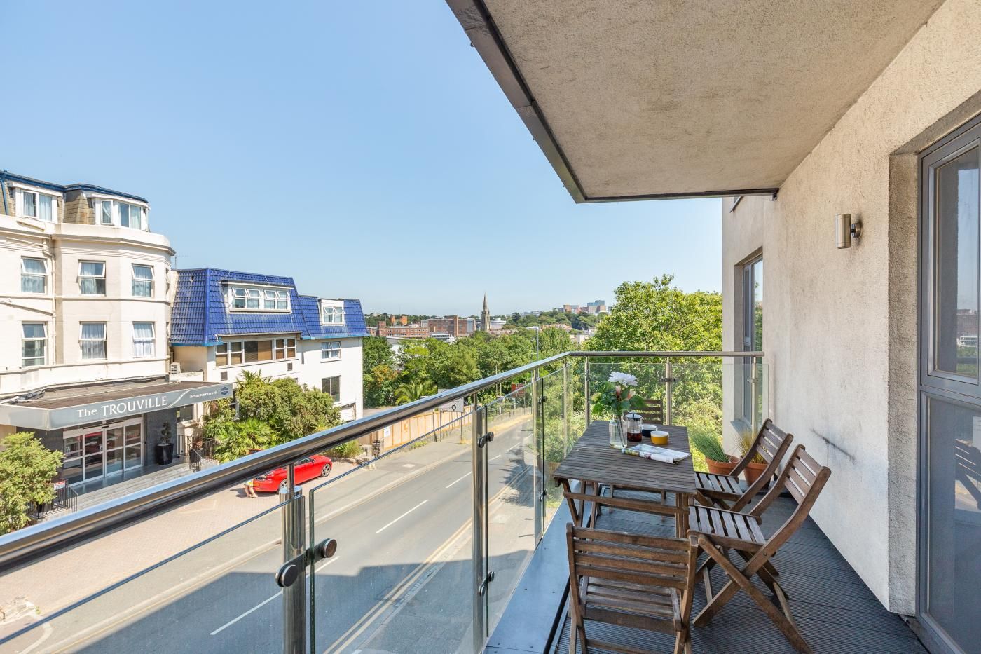 A balcony with a table and chairs, overlooking a street with buildings and a clear sky.