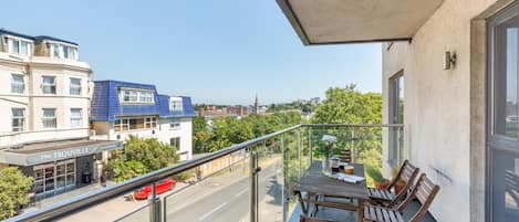 A balcony with a table and chairs, overlooking a street with buildings and a clear sky.