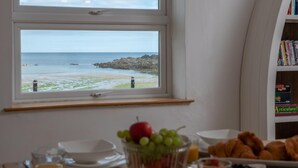 Une table de petit-déjeuner avec un panier de fruits, des croissants et une vue sur la plage par la fenêtre.