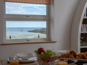 Ein Esstisch mit Obstschale, Croissants und Blick auf den Strand durch ein Fenster.